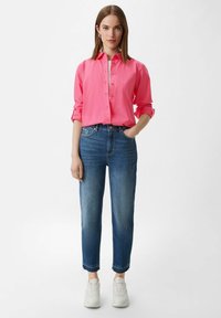 Roze overhemd met opgerolde mouwen, gecombineerd met blauwe jeans met een taps toelopende pasvorm en een rafelige zoom. Witte sneakers maken de outfit compleet.