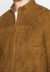Brun mockakjacka med ståkrage, dragkedjestängning och subtila sömdetaljer. Slät yta med mjuk finish.