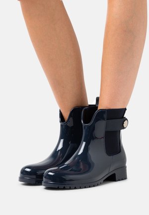 Botas tobilleras de goma azul marino con acabado brillante, paneles laterales elásticos y una correa decorativa con un botón redondo. Suela con dibujo para mayor agarre.