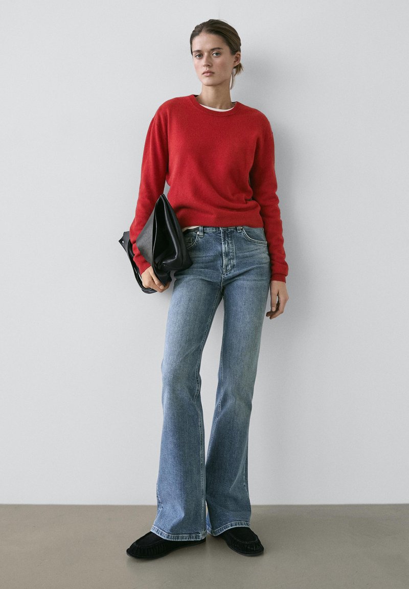 Pull en laine rouge, jean évasé bleu clair et pochette en cuir noir. La tenue présente un design simple avec des textures lisses et des couleurs vives.