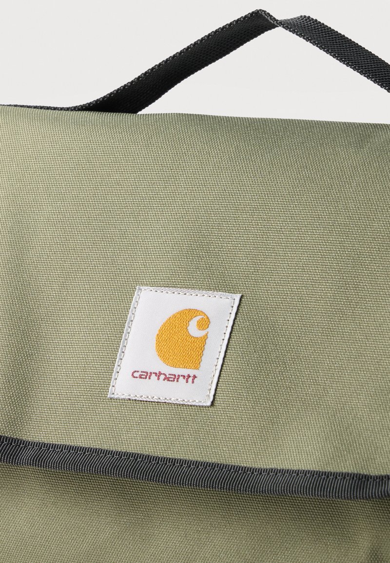 Groene stoffen rugzak met zwart handvat en witte vierkante Carhartt logopatch met een geel embleem en rode tekst op de voorklep.