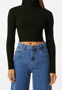 Woman wearing a black ribbed long-sleeve cropped turtleneck sweater and high-waisted blue denim jeans with hands relaxed by sides.