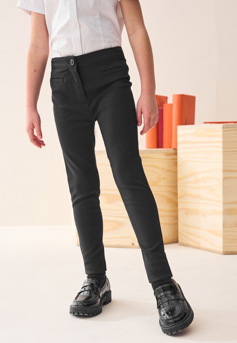 Pantaloni aderenti neri in tessuto liscio, con chiusura a bottone e tasche laterali, abbinati a scarpe nere lucide.