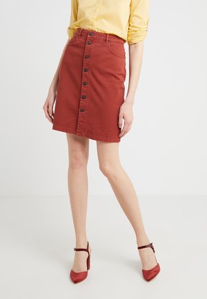 Femme portant une jupe boutonnée couleur rouille, un chemisier jaune aux manches retroussées, et des talons à bride à la cheville assortis couleur rouille.