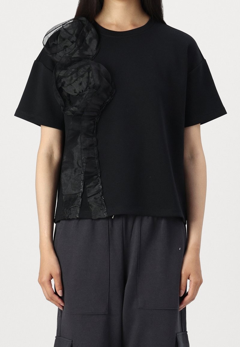 Černé tričko s krátkými rukávy, které na levé straně zdobí černý texturovaný detail z tkaniny, vytvářející vrstevnatý, prostorový efekt.