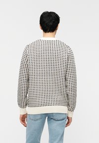 Homme portant un pull à motifs blancs et noirs et un jean bleu clair, debout de dos devant un fond blanc uni.