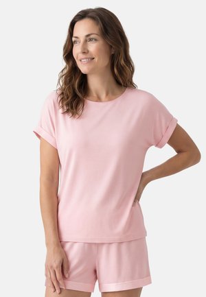 Vrouw met golvend bruin haar, gekleed in een lichtroze top met korte mouwen en bijpassende short, staand met één hand op haar heup en glimlachend.
