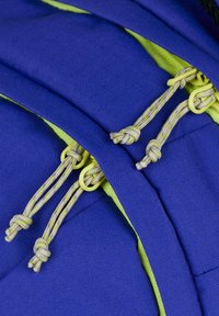 Rucksack aus blauen und grünen Stoffen mit gelben Beschlägen, strukturierten Oberflächen und Kordelzugverschlüssen mit Schlaufenverbindern.