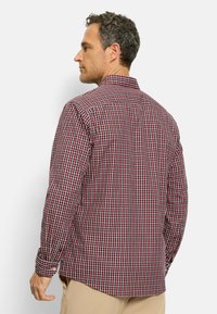 Mann trägt ein rot-schwarz-weiß kariertes Langarmhemd und beige Hose, blickt nach links mit einem neutralen Gesichtsausdruck vor weißem Hintergrund.