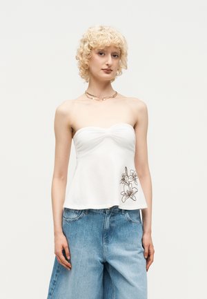 Personne blonde aux cheveux courts et bouclés porte un haut blanc sans bretelles avec un motif floral et un jean bleu clair ample, debout devant un fond uni.