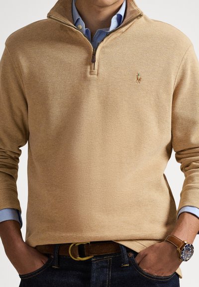 Muž v pískové svetr s krátkým zipem přes modrou košili s límečkem, džíny, hnědý kožený opasek a hnědé náramkové hodinky s námořnicky modrým ciferníkem, s rukama v kapsách.