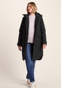 Woman walking wearing a black hooded long coat, light pink sweater, blue jeans, and white sneakers against a plain beige background.