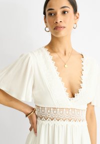 Robe blanche avec des manches courtes et bouffantes, présentant un décolleté en V profond, un bord en dentelle et un design de taille texturé. Tissu lisse, longueur genoux.