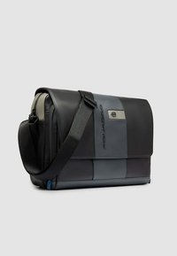 Schwarze und graue Leder-Umhängetasche mit verstellbarem Schultergurt, Klappe auf der Vorderseite und Metall-Logoschild vor einem einfarbigen Hintergrund.