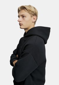 Musta huppari, jossa on iso, pehmeä ja vuorattu huppu sekä rento istuvuus. Ominaisuuksiin kuuluu leveät hihat ja ribbireunukset, sekä minimaalinen näkyvä brändäys.