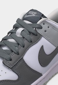 Gros plan sur une chaussure Nike blanche et grise montrant les lacets, le logo swoosh et les panneaux en cuir cousu.