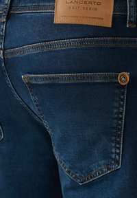 Granatowe dżinsy dzianinowe z kontrastującymi pomarańczowymi szwami, skórzaną metką marki oraz boczną kieszenią z okrągłym brązowym guzikiem.