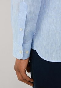 Camicia di lino azzurro chiaro con finitura testurizzata, caratterizzata da polsini con bottone e orlo curvo. Il tessuto presenta una leggera variazione di colore.