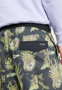 Pantalones cortos Volcom hechos de tela que presenta un fondo negro con patrones de dragones dorados y verdes; incluye una etiqueta negra en el bolsillo y una cinturilla elástica.