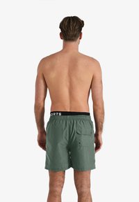 Groene zwembroek met een zwarte elastische tailleband met het label "SPORTS." Voorzien van een achterzak met een knoop en een gladde stoftextuur.