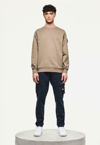Ein junger Mann steht auf einer runden Plattform, trägt einen beigefarbenen Sweatshirt, navyfarbene Cargo-Hosen mit Taschen und weiße Turnschuhe vor einem schlichten hellen Hintergrund.