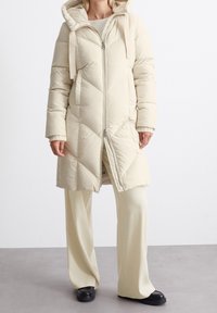 Beige vuorattu takki, jossa on huppu, vetoketju, viisto tikkauskuvio ja ribbireunukset. Päällä vaaleanbeige housut ja mustat kengät.