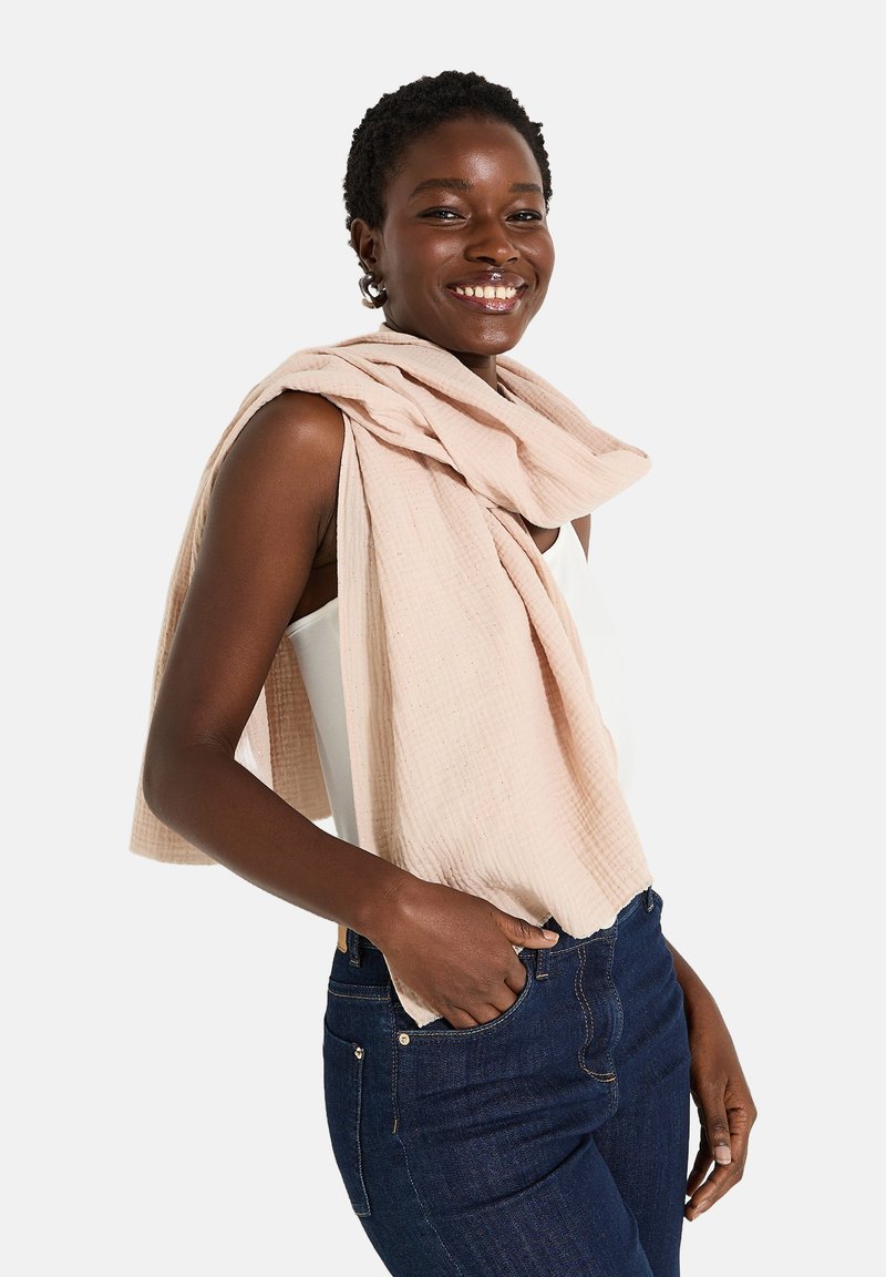 Bufanda texturizada de color rosa claro sobre los hombros, combinada con una camiseta blanca y jeans de color azul oscuro. Tejido suave con un sutil detalle de patrón.