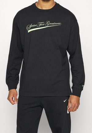 Hombre vistiendo una camisa de manga larga negra con el texto "Strive For Greatness" en verde claro y pantalones negros a juego con pequeños logotipos blancos.