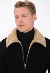 Schwarze Kunstwildlederjacke mit einem hellbraunen Lammfellkragen, mit einem Reißverschluss vorne und einem enganliegenden Rollkragenpullover darunter. Glatte Textur mit kontrastierenden Farben.