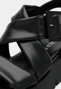 Sandale en cuir verni noir avec des larges brides croisées et une boucle rectangulaire, dotée d'une semelle épaisse et d'une texture de surface lisse.