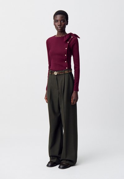 Top bordeaux a maniche lunghe con fiocco sulle spalle e bottoni dorati, abbinato a pantaloni larghi a vita alta di un verde oliva e scarpe nere.