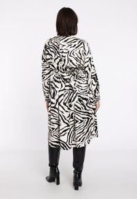 Langes Kleid mit einem schwarz-weißen Zebra-Muster, aus strukturiertem Stoff, mit gebundenem Taillengürtel und langen Ärmeln. schwarze kniehohe Stiefel.