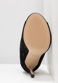 Chaussure à talon haut en daim noir reposant sur le côté, montrant une semelle beige avec une accroche ovale striée et le logo ALDO, sur fond blanc.