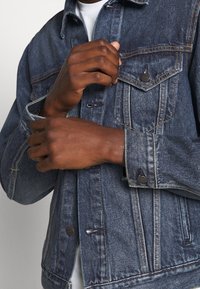 Chaqueta de denim en azul oscuro con costuras claras, bolsillos con botón y un cuello clásico. Presenta una textura ligeramente áspera y un ajuste holgado.