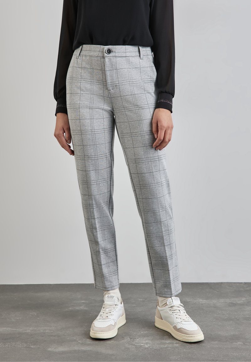 Personne portant un pantalon slim à carreaux gris, des baskets blanches et un haut noir à manches longues, debout sur un sol gris.