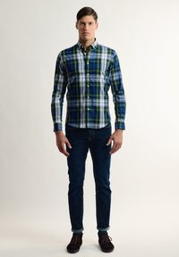 Camisa a cuadros en azul, verde y blanco. Presenta mangas largas, un cuello y un bolsillo en el pecho. Combinada con jeans oscuros y zapatos marrones.