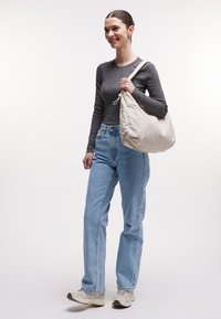 Sac fourre-tout en tissu beige clair, forme ronde, avec un haut froncé et un cordon de serrage. Associé à une chemise à manches longues grise et un jean bleu, porté avec des baskets.