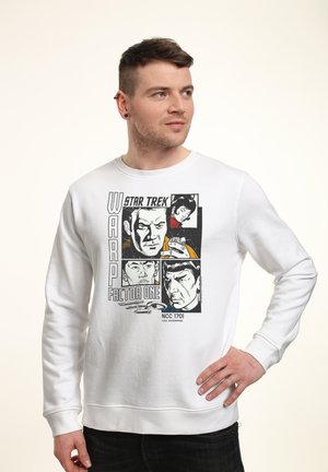 Man draagt een wit sweatshirt met Star Trek-stripstijl gezichten en tekst, staat met hand op heup, kijkt naar rechts.
