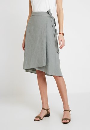 Mujer con una falda cruzada gris claro atada a la cintura con un lazo, combinada con sandalias de tacón grueso en color verde oliva y punta abierta.