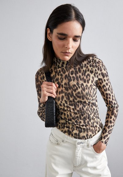 Langermige top met luipaardprint in bruin- en tan-tinten, gecombineerd met lichtgekleurde jeans. Het model houdt een gestructureerde zwarte tasband vast.