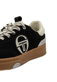Sneaker in suede nera con logo bianco, chiusura con lacci testurizzati e suola in gomma marrone. Presenta un dettaglio sul tallone bianco a contrasto.