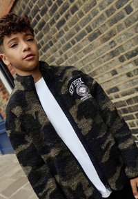 Többszínű, katonai álcás mintázatú fleece zipzáras dzseki, fekete és olíva árnyalatokkal, fehér bélésével, és hímzett "NEW YORK" logóval a mellkasán.