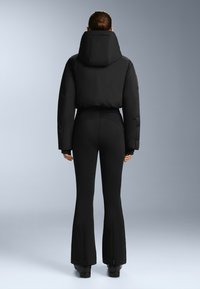 Czarna krótka kurtka puchowa z dużym kapturem, zestawiona z dopasowanymi czarnymi spodniami dzwonami z zamkami przy kostkach. Gładka tekstura, minimalistyczny design.