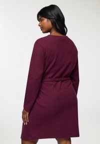 Abito bordeaux in morbido tessuto con maniche lunghe, vita cintura e scollo rotondo. Il design presenta una texture semplice e liscia.