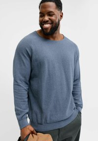 Lyseblå teksturert genser med vid halsutringning og ribbestrikkede mansjetter, kombinert med mørke bukser. Modellen holder en beige jakke.