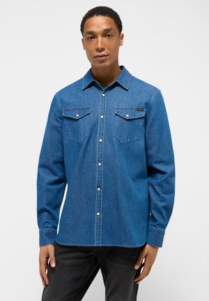 Camisa de mezclilla azul con cuello, mangas largas y dos bolsillos en el pecho. Presenta botones plateados y un dobladillo ligeramente curvado.