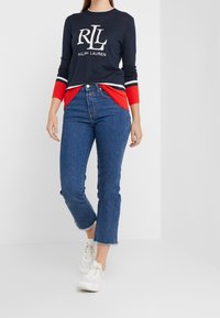 Kobieta ubrana w granatowy i czerwony sweter Ralph Lauren, jeansy z wysokim stanem i ściętymi nogawkami w kolorze niebieskim oraz białe trampki, stojąca na białej podłodze.