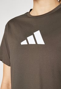 Tamno smeđi majica s kratkim rukavima od pamuka s bijelim logom od tri pruge na prsima. Dizajn s okruglim izrezom. Glatka tekstura.