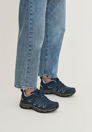 Persoon draagt lichtblauwe spijkerbroek en donkerblauwe sportschoenen met veters en lichtblauwe accenten, staand op een lichte vloer.