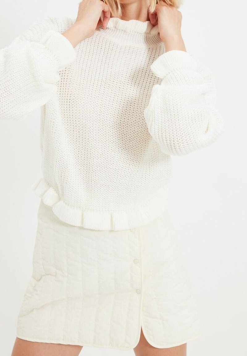 Weißer Strickpullover mit Rüschenausschnitt und -ärmeln, kombiniert mit einem strukturierten weißen Rock mit Knöpfen und einem Schlitz vorne.